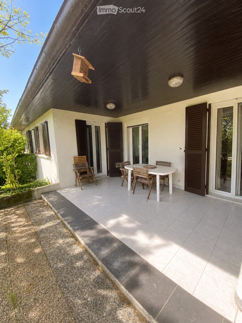 Balcony with a white table and four wooden chairs, surrounded by glass doors and wooden shutters, with a wooden birdhouse hanging from the ceiling.