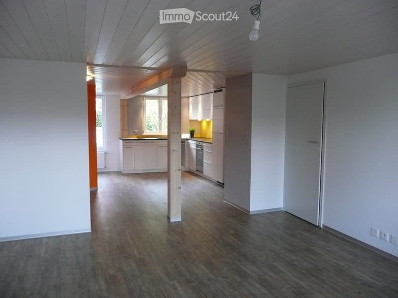 Empty room with hardwood floor, wooden beams, kitchen area with windows, white cabinets, yellow countertop, appliances, white walls