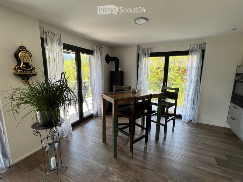Bright dining area with wooden table and chairs, large windows with white curtains, potted plant, and decorative wall clock.