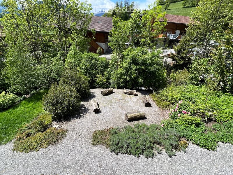 Garden area with gravel pathway, wooden logs, shrubs, and flowers, surrounded by trees and houses in the background.