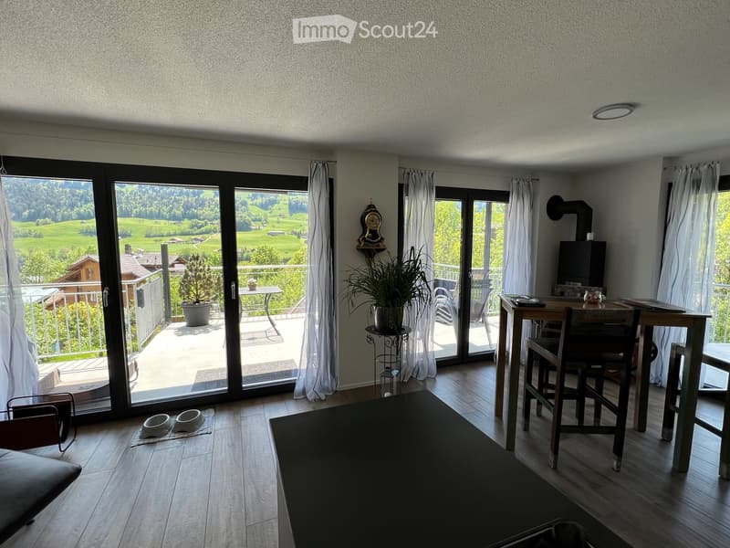 Modern living room with large windows offering a scenic view, featuring a sofa, dining table, chairs, a plant, and a clock on the wall.
