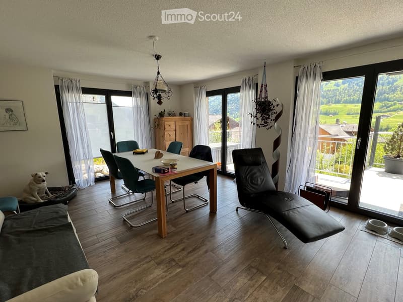 Bright living and dining area with wooden flooring, large windows offering a scenic view, a sofa, a dining table with chairs, a dog and its bed, a leather chair, a small cupboard, and decorative items.