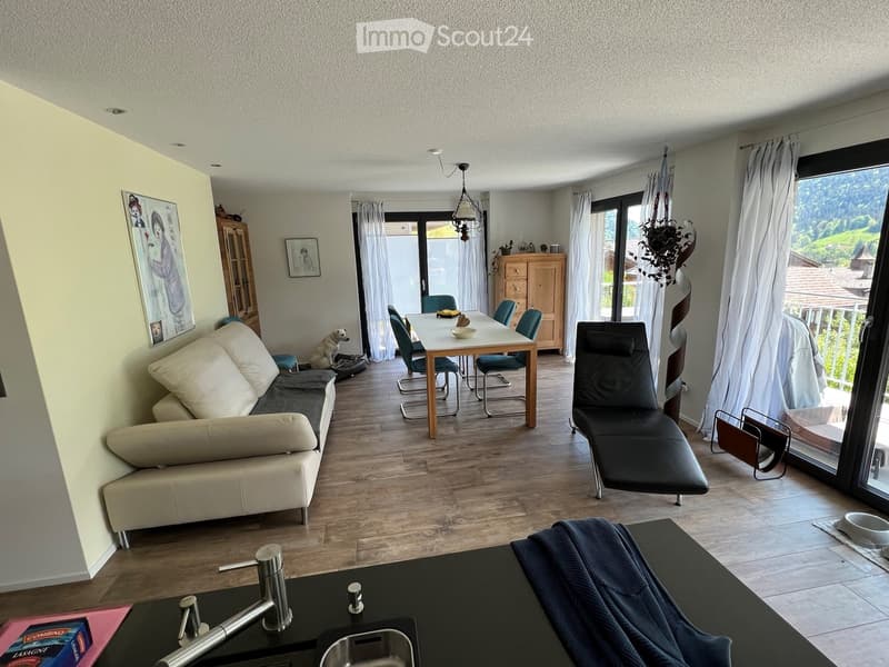 Open-plan living and dining area with a sofa, dining table, chairs, dog, and a view through large windows.