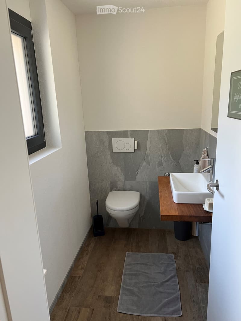 Modern bathroom with a toilet, sink, wooden vanity, and grey tiled accent wall. Includes a grey bathroom rug and a small window.