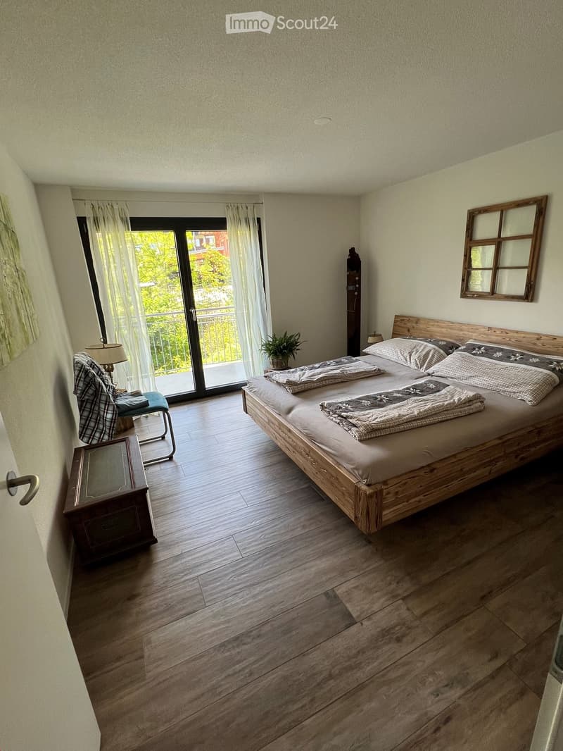 Cozy bedroom with wooden bed, nightstand, chair, lamp, and a view through large windows.