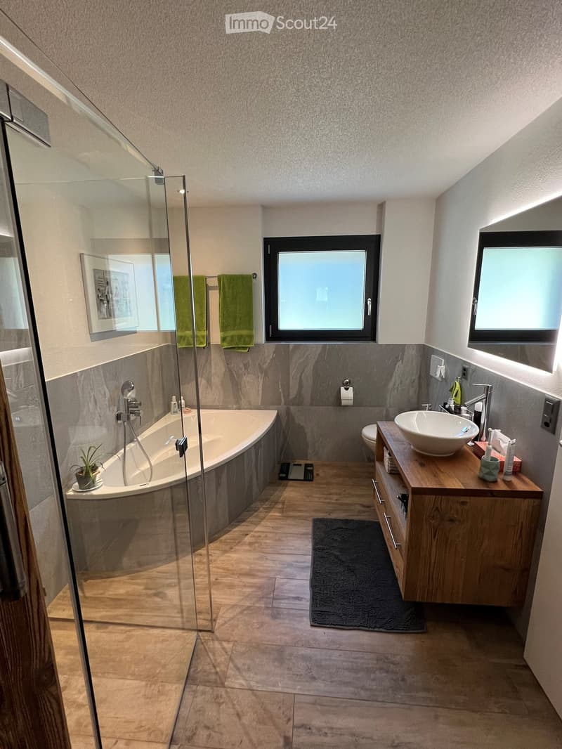 Modern bathroom with a bathtub, sink, toilet, and wooden cabinet. Features a glass shower enclosure, tiled walls, and a small plant on the floor.
