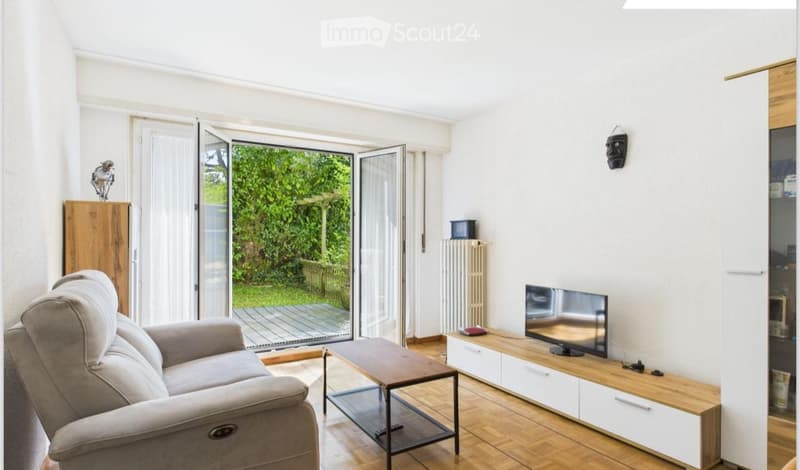 Bright living room with large sliding glass doors opening to a garden, grey sofa, wooden coffee table, TV on a white and wood stand, radiator, and a wooden cabinet with decorative items.