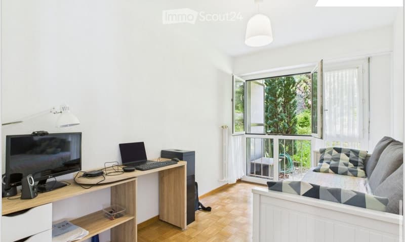 A modern, well-lit room with a sofa, a desk with a laptop, and a large window with a view of greenery.