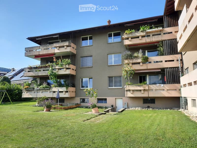 Multi-story residential building with balconies, greenery, and a well-maintained lawn in front.