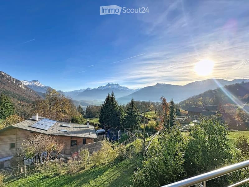 A wooden house with solar panels on the roof, surrounded by trees and a scenic mountain view in the background.