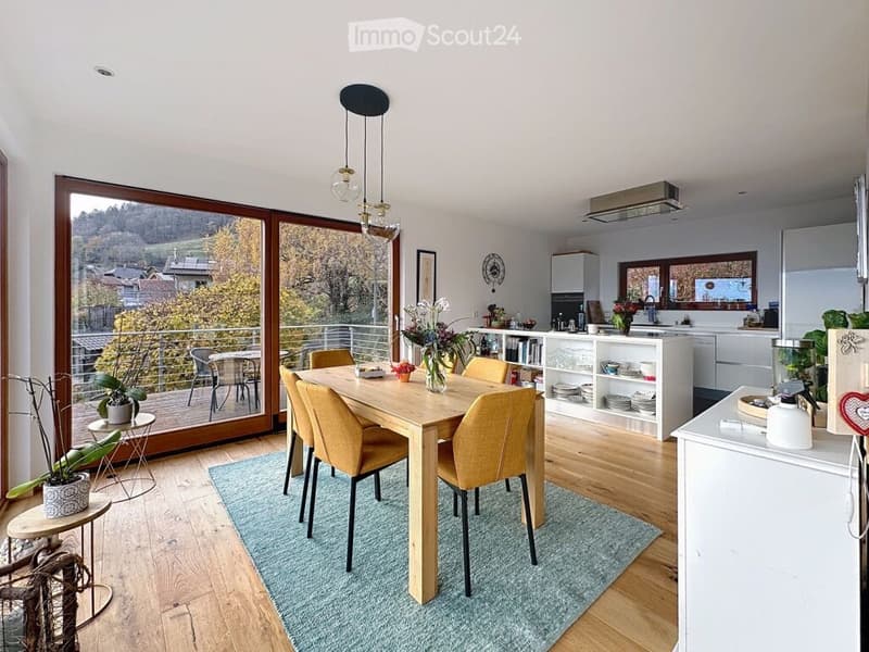 Bright, open-plan dining and kitchen area with wooden flooring, large windows offering a scenic view, a dining table with six chairs, modern kitchen cabinets, and decorative elements like potted plants and a vase.