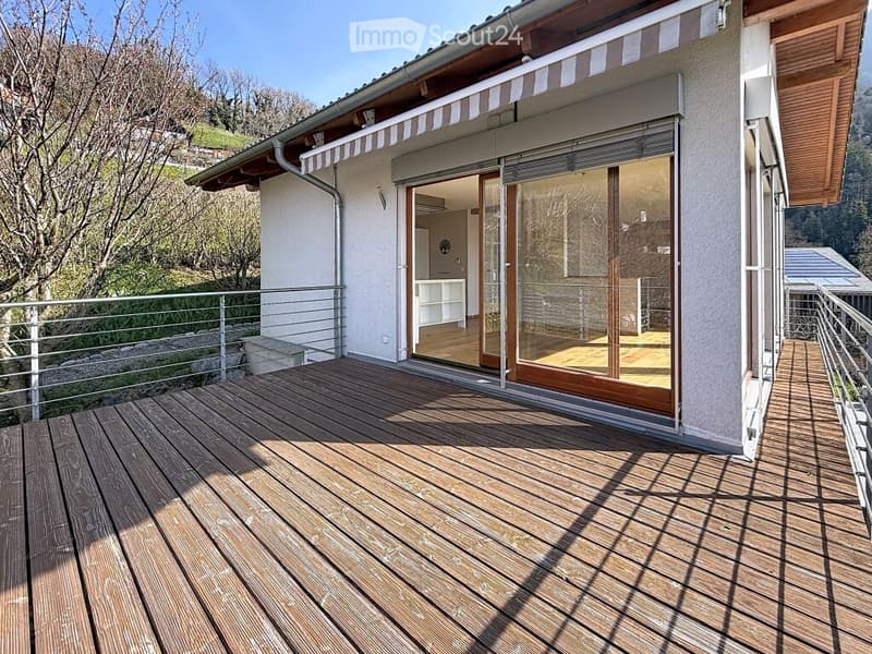 Wooden deck with metal railing, large sliding glass doors leading to interior, view of greenery and mountains.