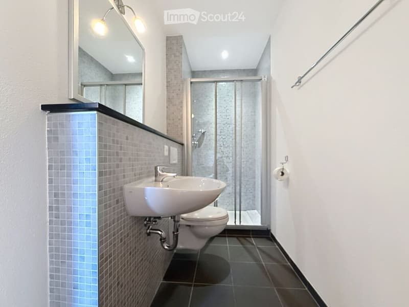 Modern bathroom with a white sink, toilet, and glass-enclosed shower. Features a large mirror with built-in lighting and tiled walls.