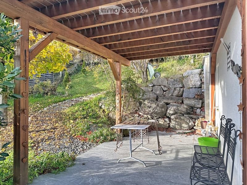 Wooden pergola-covered patio with a stone wall backdrop, small table, chairs, and surrounding garden area with plants and trees.