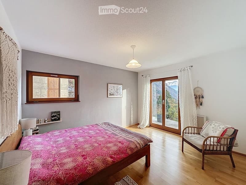 Bright bedroom with wooden flooring, large window, and sliding glass door leading to a balcony with mountain view. Features a double bed with pink bedding, a wicker chair, a table lamp, and a picture frame on the wall.
