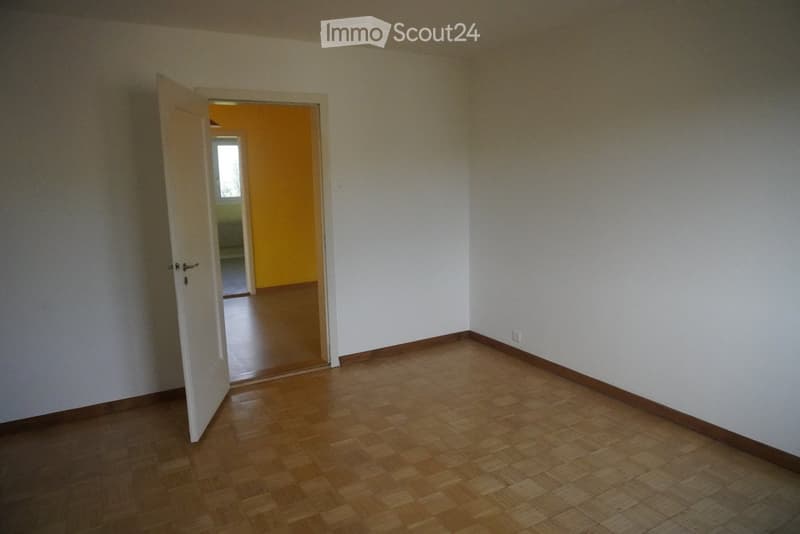 Empty room with wooden flooring and white walls, featuring an open doorway leading to another room with yellow walls and a visible window.