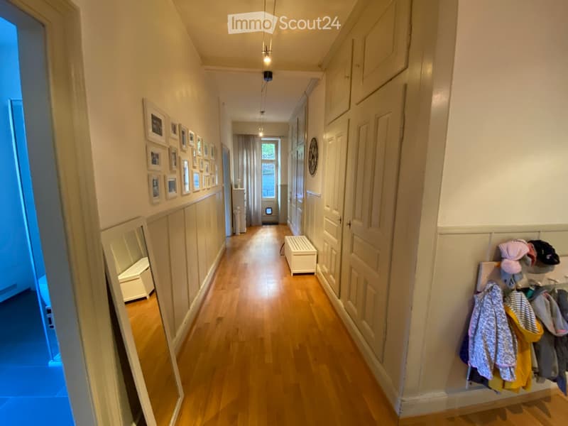 A long, narrow hallway with wooden flooring, white walls, and several doors. There are framed pictures on one wall and a coat rack with various items on the other side.