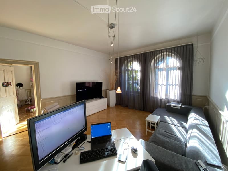 A modern living room with a grey sofa, a white coffee table, a laptop, a large screen, and a vase with dried plants. The room features large arched windows with dark curtains and wooden flooring.