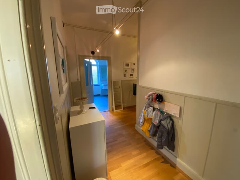 A narrow hallway with wooden flooring, white walls, and a coat rack holding several jackets. A small table with a cake and a phone is visible on the left side, and a doorway leads to another room.