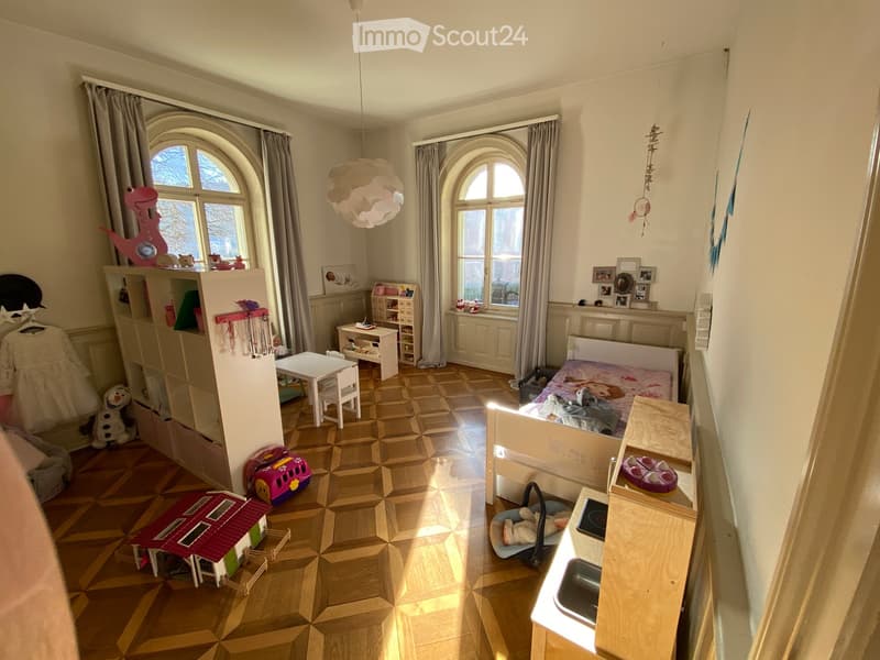 A well-lit bedroom with a bed, a small table and chairs, shelves filled with toys and books, and a large window with curtains. The room features a patterned wooden floor and decorative wall items.