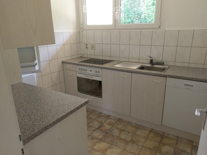 Modern kitchen with light wood cabinets, granite countertops, built-in oven, sink, and dishwasher. The space features tiled walls and a window with a view of greenery.
