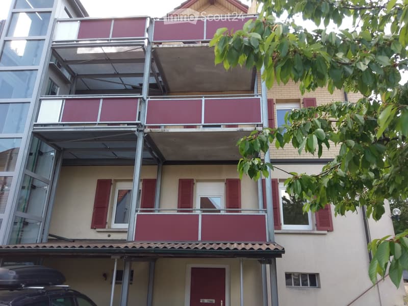 Multi-story residential building with balconies, red shutters, and a parked car in the foreground.