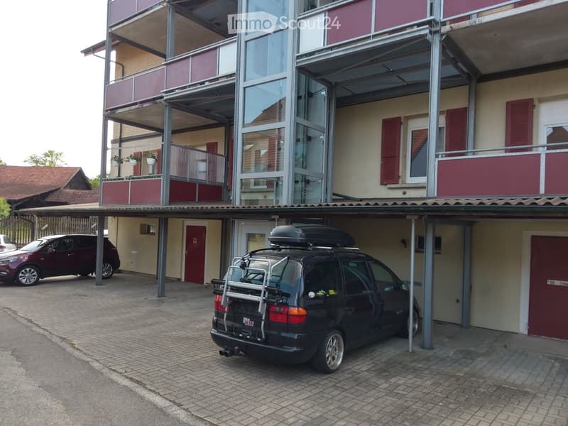 Multi-story building with a covered parking area, featuring a black SUV with a roof rack parked inside.