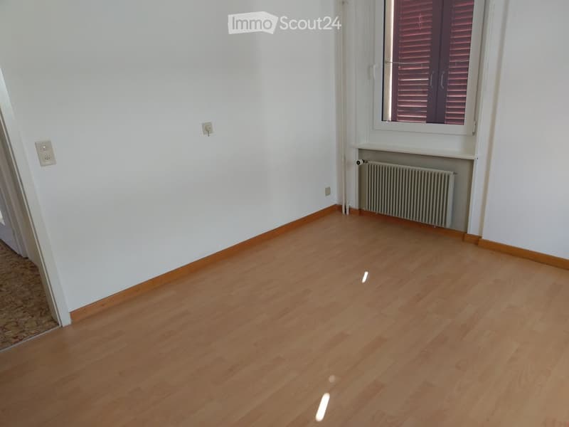 Empty room with light wooden flooring, white walls, and a window with blinds. There is a radiator below the window and electrical outlets on the walls.