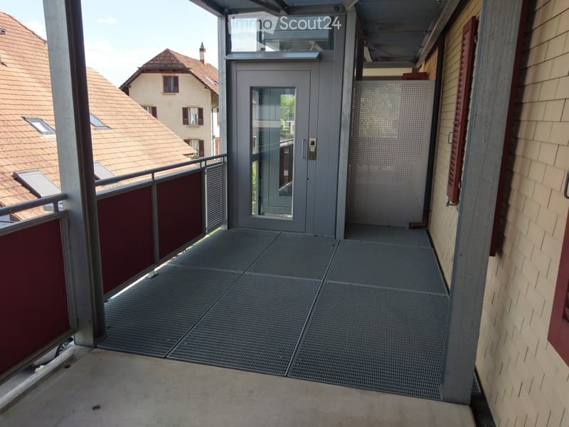 Balcony with metal floor, glass elevator door, red paneling, and view of neighboring houses.