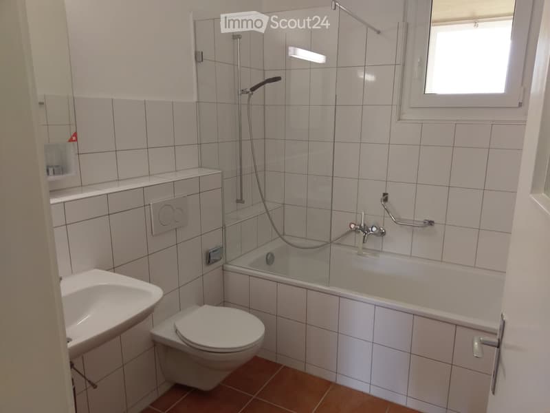 Modern bathroom with white tiled walls, a bathtub with a glass shower enclosure, a toilet, a sink, and a window providing natural light.