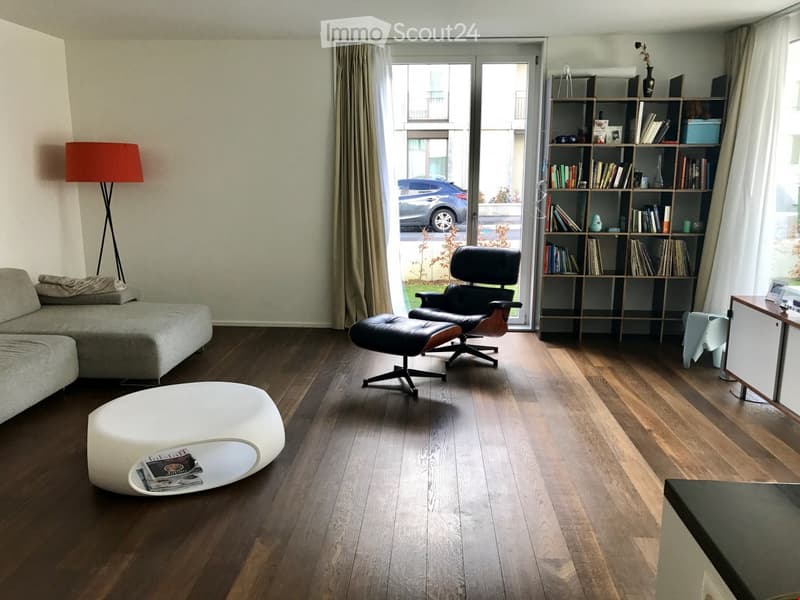 Living room with wooden floor, gray couch, white ottoman, black leather chairs, bookshelf, glass door, red lamp, and a view of a car outside.