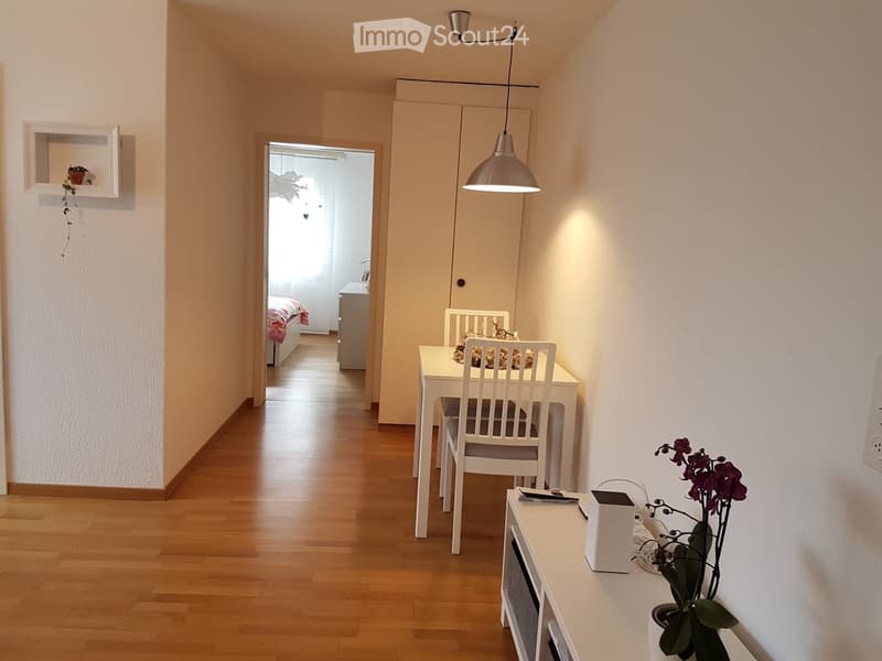 A modern hallway with a small dining area featuring a white table and chairs, a flower pot on a side table, and a visible bedroom in the background with a bed and nightstand.