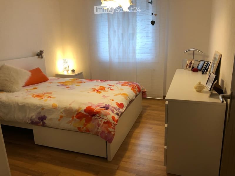 Cozy bedroom with a large bed, colorful floral duvet, nightstand with lamp, and a dresser with picture frames and decorative items.