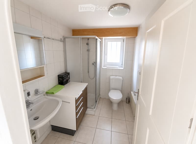 Modern bathroom with white tiles, a sink with a mirror, a toilet, a glass-enclosed shower, and a window for natural light.
