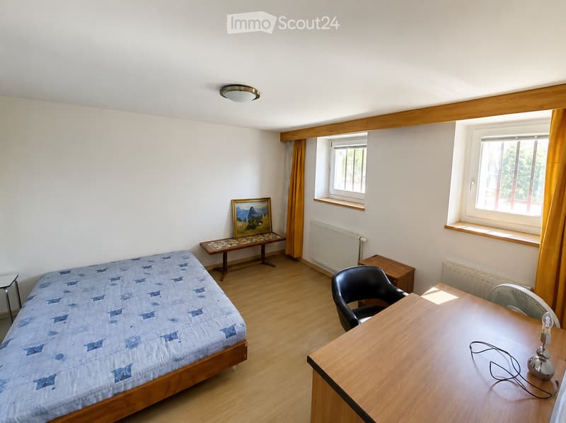 A furnished bedroom with a single bed, a desk with a chair, a small table with a picture frame, and a lamp on the desk.
