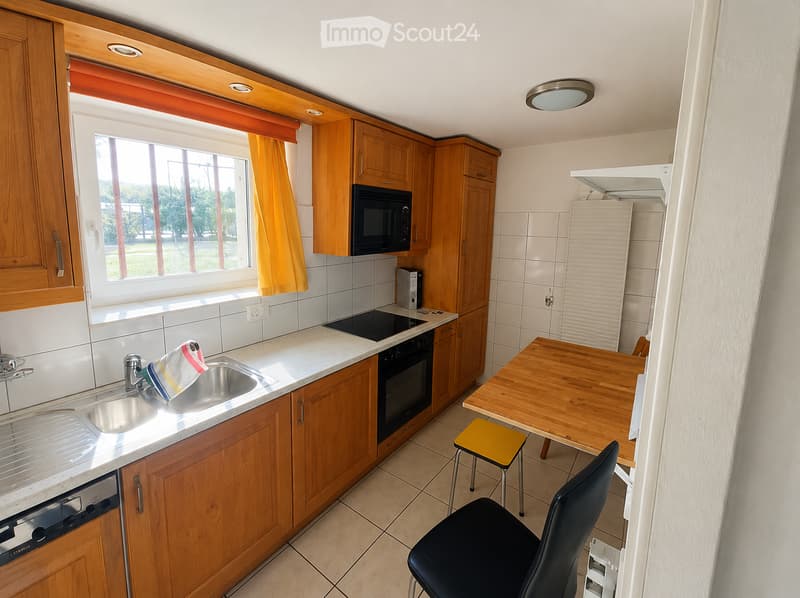 Modern kitchen with wooden cabinets, white tiled walls, and a marble countertop. Features include a sink, oven, microwave, dishwasher, refrigerator, and a small dining area with a table and chairs.