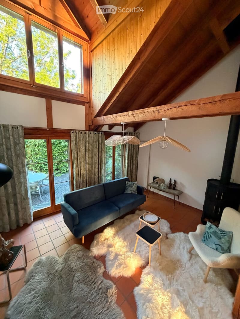 A cozy living room with wooden beams and exposed wooden ceiling, large windows with wooden frames, and a sliding glass door leading to a patio. The room features a blue sofa, a white armchair, a small wooden table with a bowl, a wooden shelf with decorati