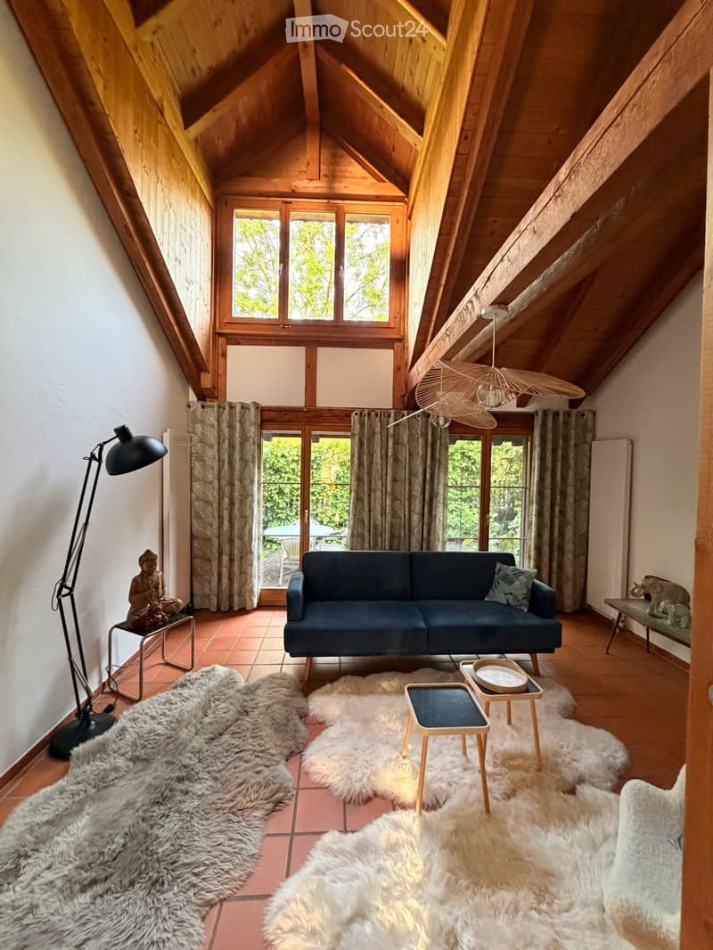 Living room with wooden beams, large windows, a dark blue sofa, wooden stools, a floor lamp, and fur rugs.