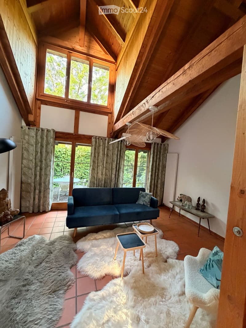 A cozy living room with exposed wooden beams, large windows, and a blue sofa. The room features multiple fur rugs, small wooden stools, a side table, and decorative items on a shelf.