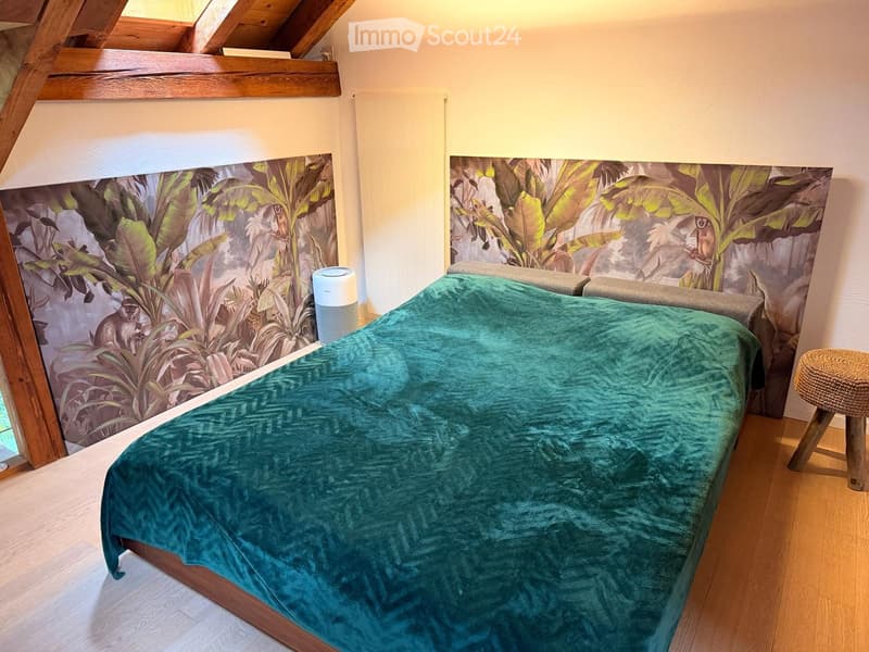 Cozy attic bedroom with wooden beams, a bed covered in a teal duvet, a small stool, and a speaker on a shelf. The walls feature a large, leafy mural.