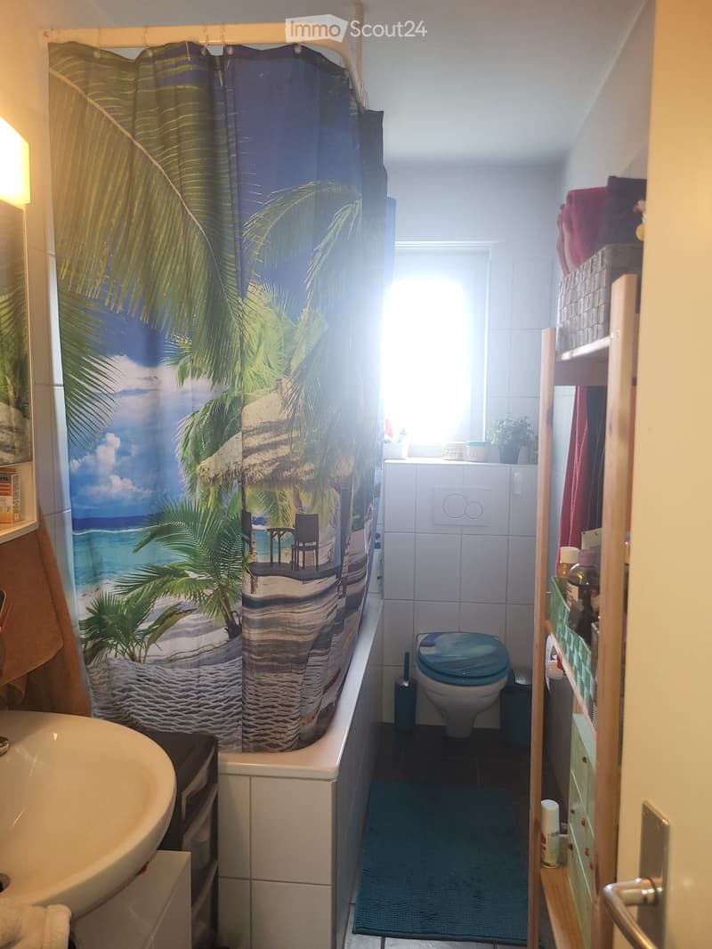 Cozy bathroom with a bathtub, toilet, sink, and a wooden shelf holding towels and toiletries. The shower curtain features a beach theme with palm trees and a beach scene.