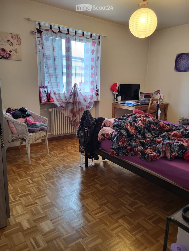 Cozy bedroom with wooden flooring, a bed with colorful bedding, a wicker chair, a desk with a computer, and a window with floral curtains.