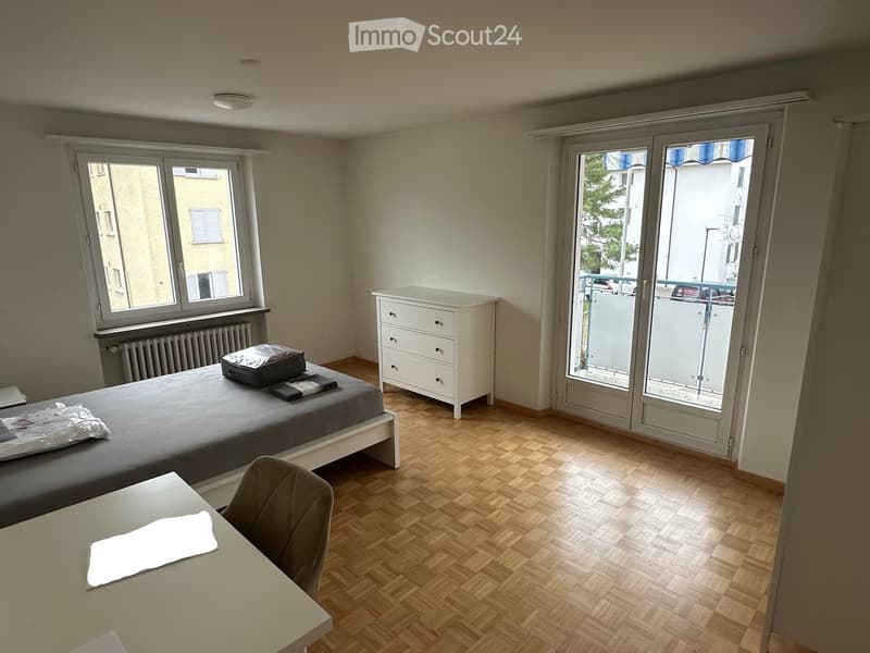 A furnished room with a bed, a chair, a table, and a drawer unit. The room has wooden flooring and large windows, one of which opens to a balcony.