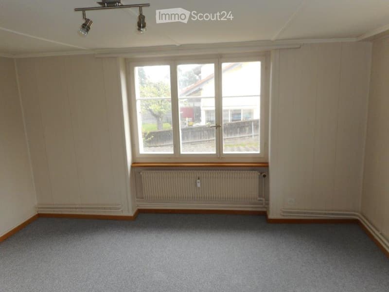 Empty room with beige walls, grey carpet, large window with wooden frame, radiator, and ceiling spotlights.