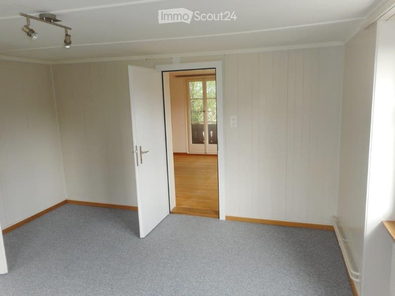 Empty room with grey carpet, white walls, and wooden baseboards. An open door leads to another room with wooden flooring and large windows.
