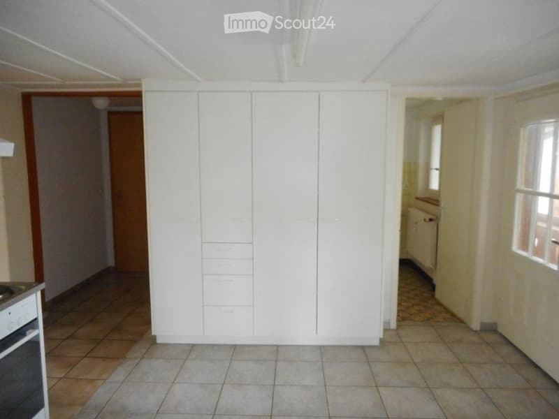 Empty room with tiled flooring, white built-in cabinetry, and adjacent kitchen area visible through an open doorway.