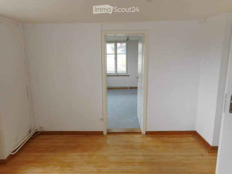 Empty room with wooden flooring and white walls, featuring a doorway leading to another room with a window and carpeted floor.