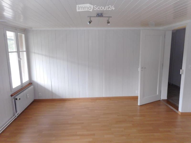 Empty room with wooden flooring, white paneled walls, and a single window with a radiator below it. The room has a white door leading to another space.