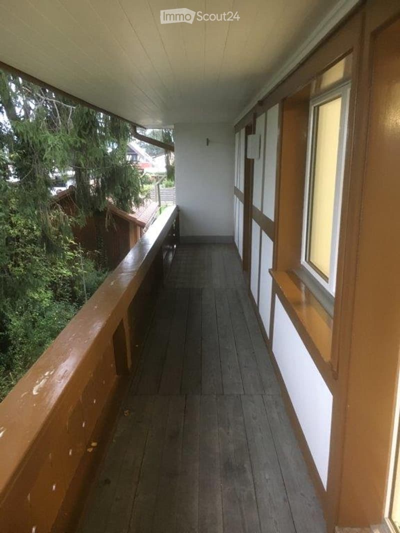 Wooden balcony with white walls, wooden railing, and large windows offering a view of greenery outside.