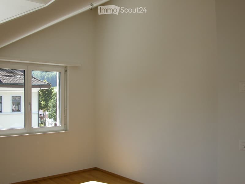 Empty room with light-colored walls and wooden flooring, featuring a large window with a view of neighboring buildings and greenery.